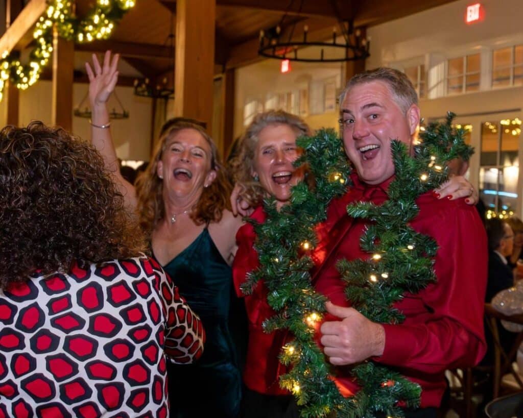 Attendees celebrating at the 2023 Holiday Gala, dressed festively with holiday lights and garland in the background, showing joy and laughter.