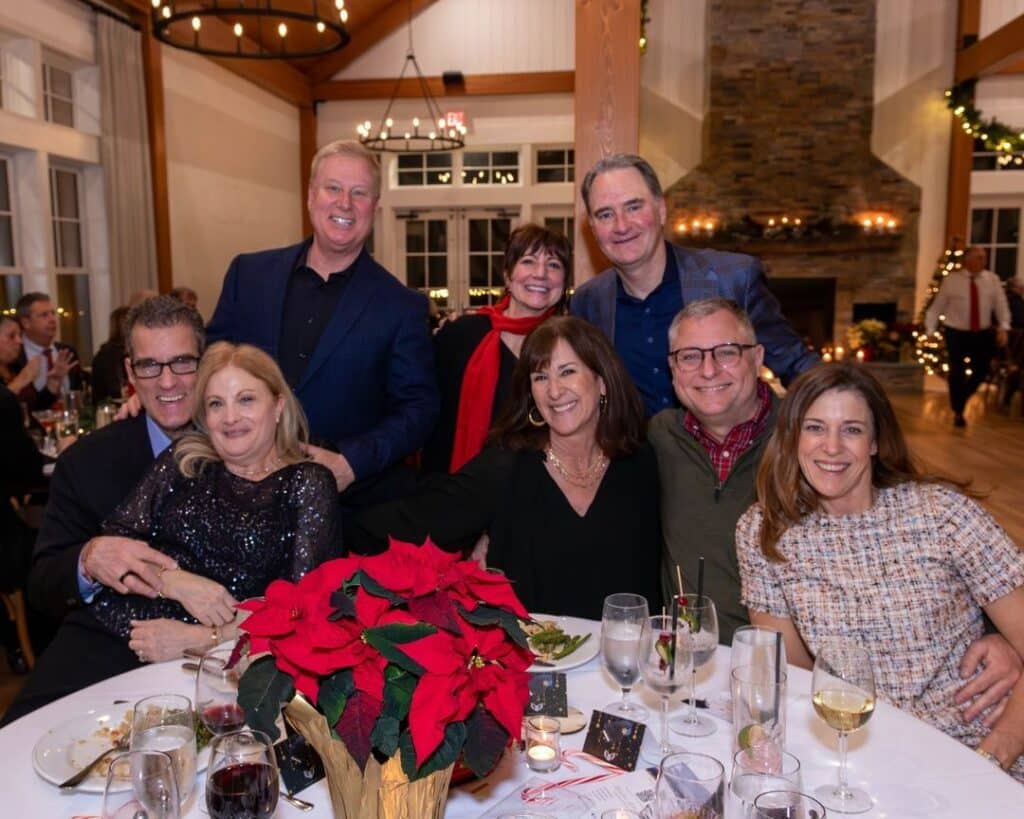 A group of smiling friends gathered around a table at the 2023 Holiday Gala at Lakehouse, with holiday decorations in the background.